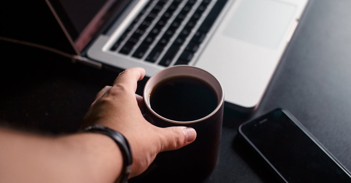 A hand reaching for a coffee mug next to a laptop streaming content on a dark desk